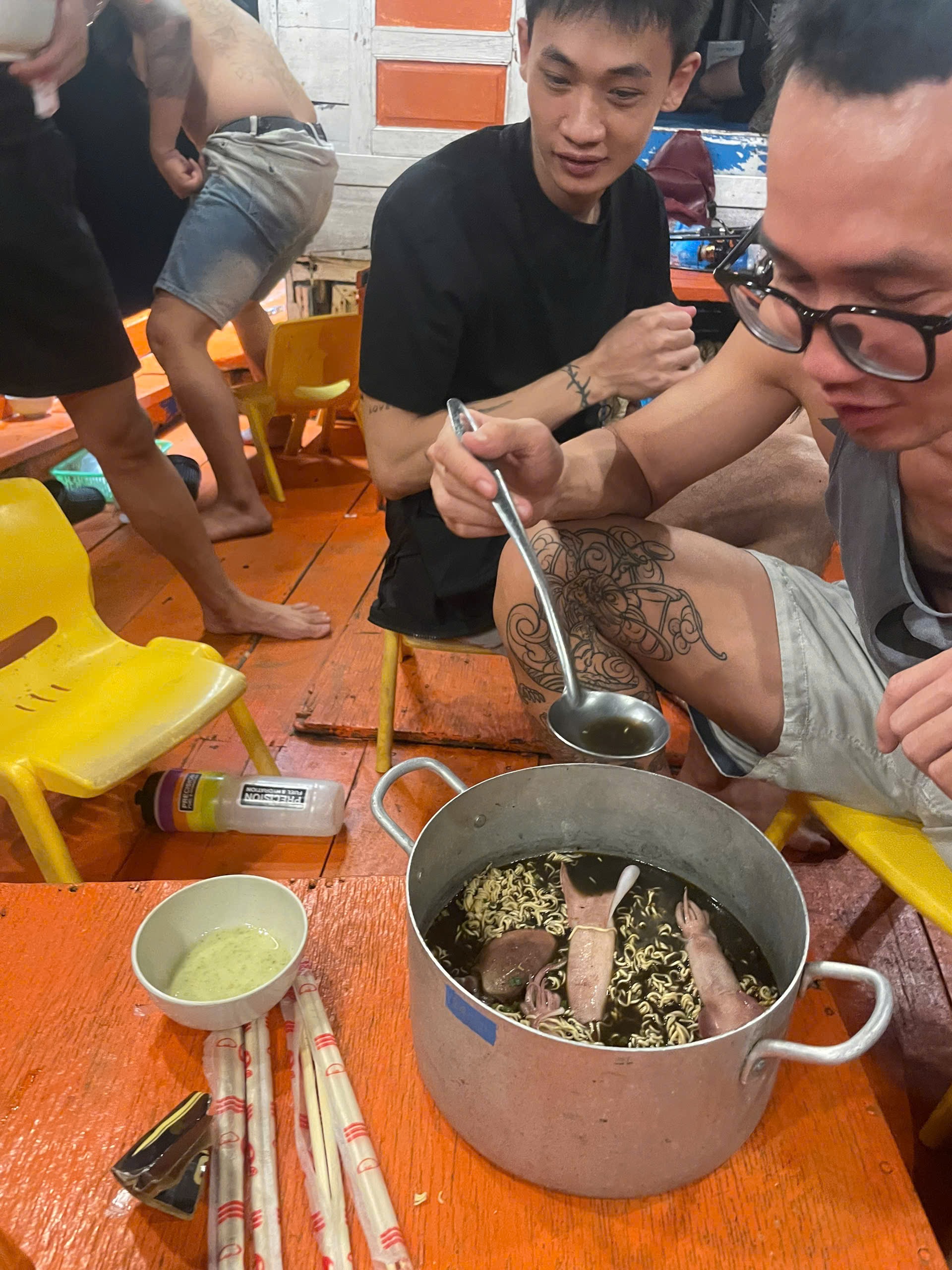 船の甲板で、参加者が小さな椅子に座り、イカ入りのインスタントラーメンの鍋を囲んでいる